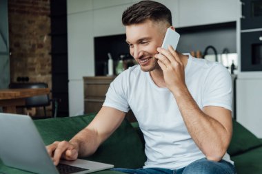 Akıllı telefondan konuşan ve evde dizüstü bilgisayar kullanan gülümseyen tele-işçinin seçici odağı 