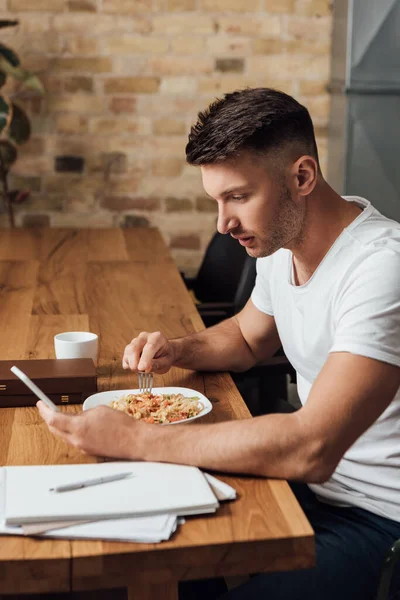 台所のテーブルの上で紙の近くの麺を食べながら スマートフォンを使用して男の側面図 — ストック写真