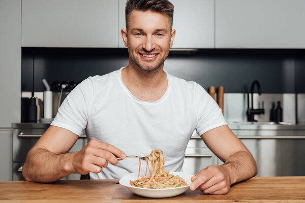 Handsome man smiling at camera while eating noodles in kitchen 