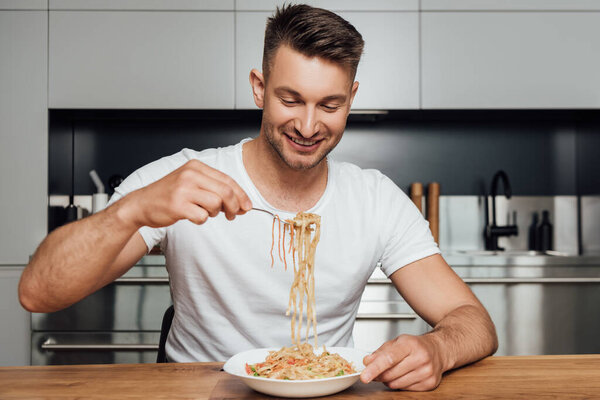 Handsome man smiling while holding fork with noodles at table in kitchen 