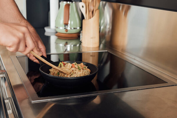 Cropped view of man holding wooden spatula while preparing noodles in frying pan in kitchen 