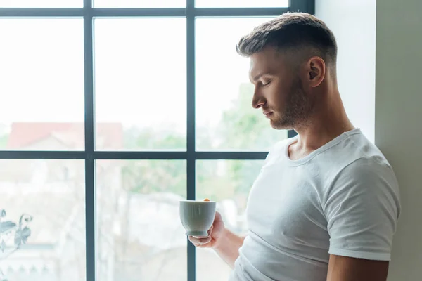 站在家里靠窗的时候拿着一杯咖啡的英俊男人的侧视图 — 图库照片