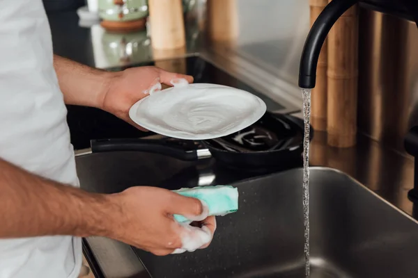 Cropped view of man cleaning plate with sponge in foam in kitchen