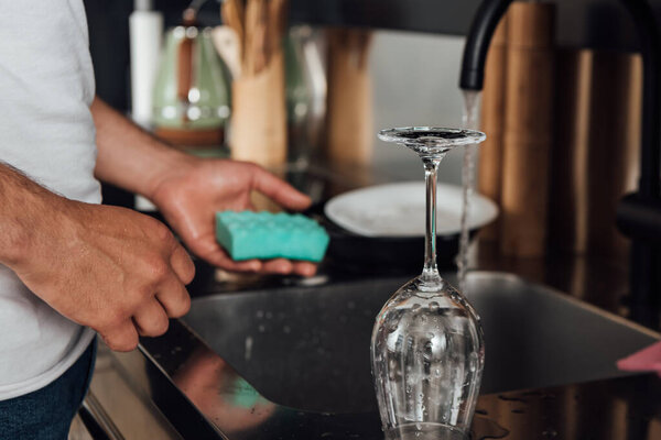 Cropped view of man holding sponge near wet wine glass and kitchen sink 