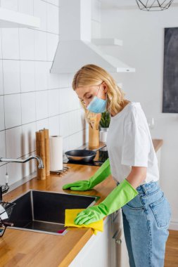 Tıbbi maskeli ve lastik eldivenli genç bir kadın mutfakta ev işi yapıyor. 