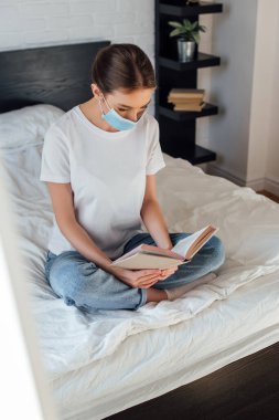 Tıp maskeli genç kadının yatakta kitap okuduğu seçmeli bir odak noktası. 
