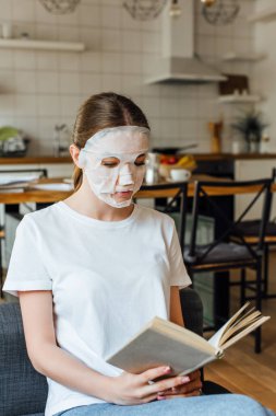 Yüzü maskeli bir kızın evdeki kanepede kitap okumasının seçici odağı. 