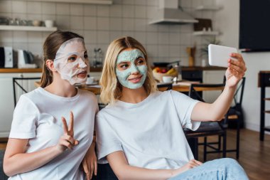 Yüz maskeleri takmış kız kardeşler evde akıllı telefonlarıyla selfie çekerken barış jestleri yapıyor ve gülümsüyorlar.
