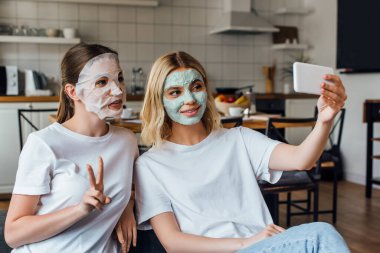 Sisters in face masks showing peace gesture and smiling while taking selfie with smartphone at home clipart