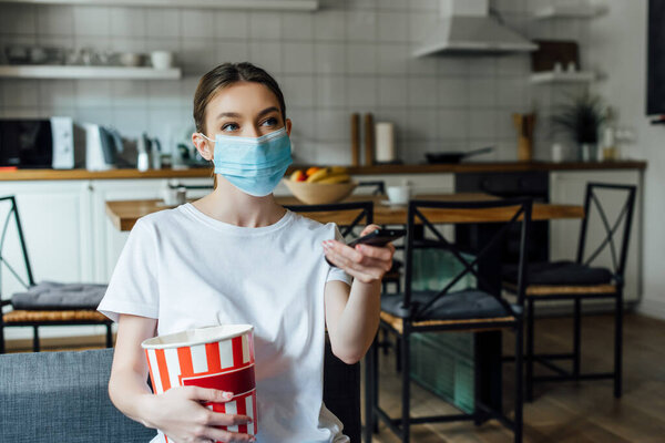 Girl in medical mask holding popcorn while watching movie on couch 