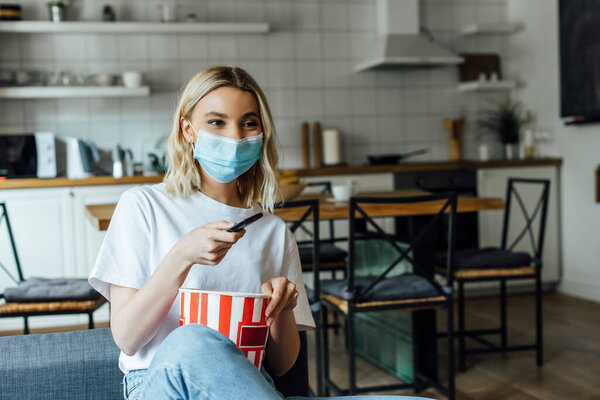 Blonde girl in medical mask holding bucket of popcorn and remote controller on sofa 