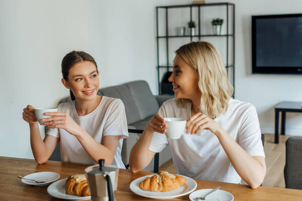 Selective focus of smiling girls drinking coffee during breakfast in kitchen 