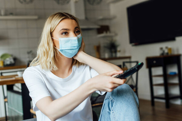 Selective focus of blonde woman in medical mask watching tv at home 