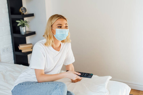 Blonde girl in medical mask holding remote controller on bed 