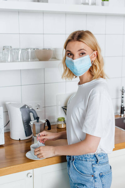 Side view of woman in medical mask looking at camera while mixing coffee in kitchen 