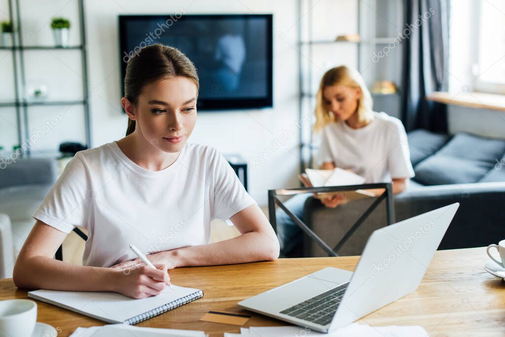 selective focus of woman looking at laptop while writing in notebook near sister, online study concept 