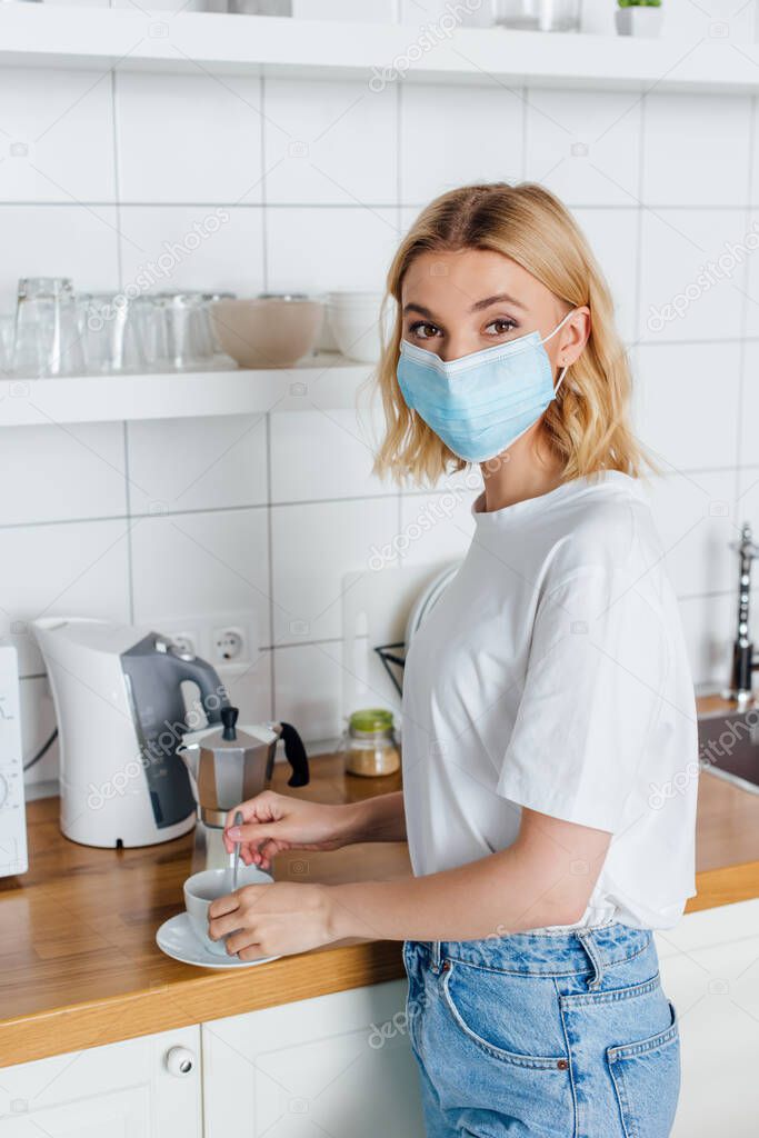 Side view of woman in medical mask looking at camera while mixing coffee in kitchen 
