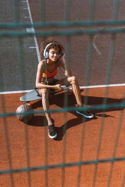 Young african-american woman in sportswear listening to music in headphones and sitting on longboard at sports ground — Stock Photo