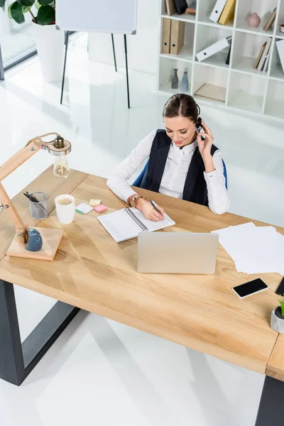 Empresária com fone de ouvido escrevendo notas — Fotografia de Stock