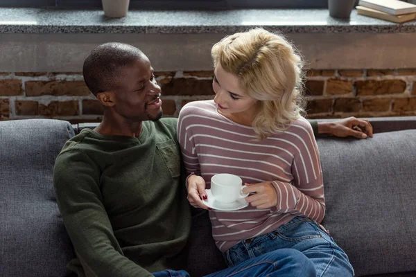 Smiling african american man looking at caucasian girlfriend with cup of hot drink in hands while resting on sofa at home — Stock Photo