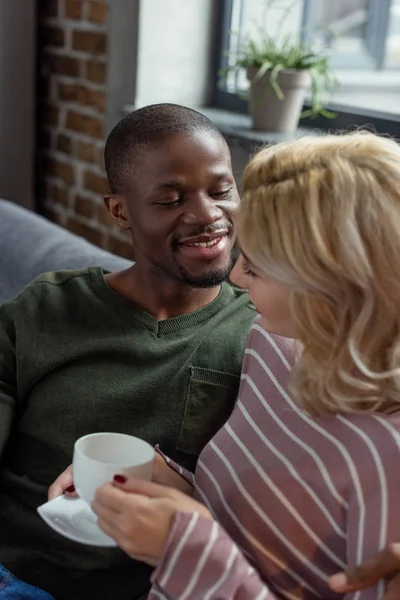 Uomo afroamericano sorridente guardando la ragazza caucasica con una tazza di bevanda calda in mano mentre riposa sul divano di casa — Foto stock