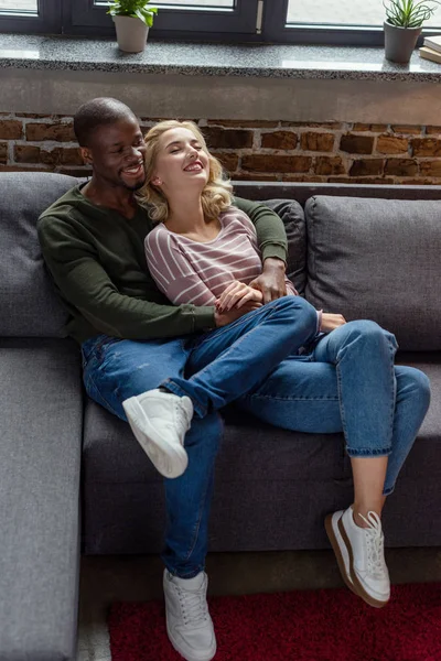 Vue grand angle de heureux homme afro-américain étreignant petite amie tout en se reposant sur le canapé ensemble — Photo de stock