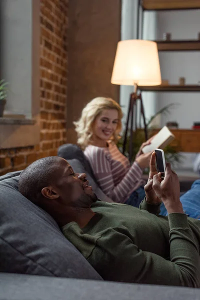 Foyer sélectif de l'homme afro-américain en utilisant un smartphone tandis que la petite amie caucasienne lecture livre sur canapé à la maison — Photo de stock