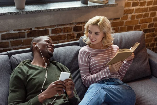 Sorrindo afro-americano homem ouvindo música enquanto a namorada caucasiana ler livro no sofá em casa — Fotografia de Stock