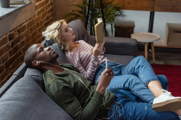 Afro-américain homme écouter de la musique tandis que caucasien copine lecture livre sur canapé à la maison — Photo de stock