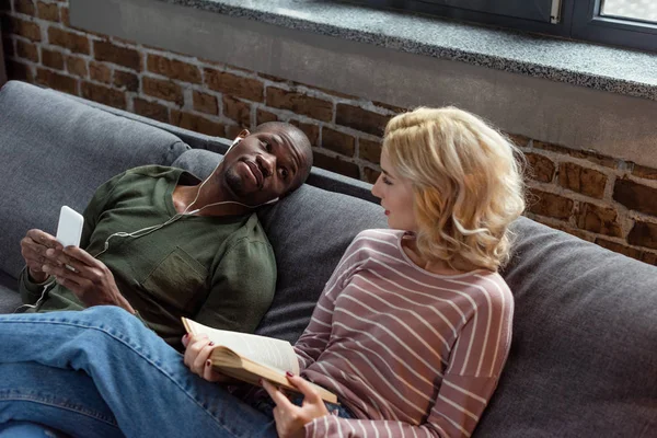 Africano americano hombre escuchar música mientras caucásico novia lectura libro en sofá en casa - foto de stock