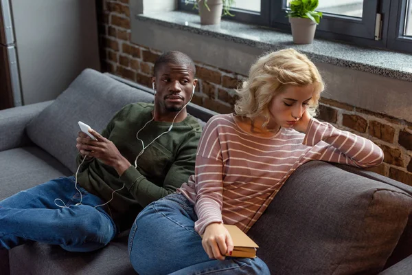 Africano americano hombre en auriculares mirando triste novia con libro en sofá en casa — Stock Photo
