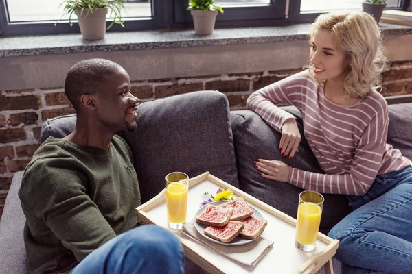 Retrato de pareja joven multicultural sentada en el sofá mientras desayunan juntos en casa - foto de stock