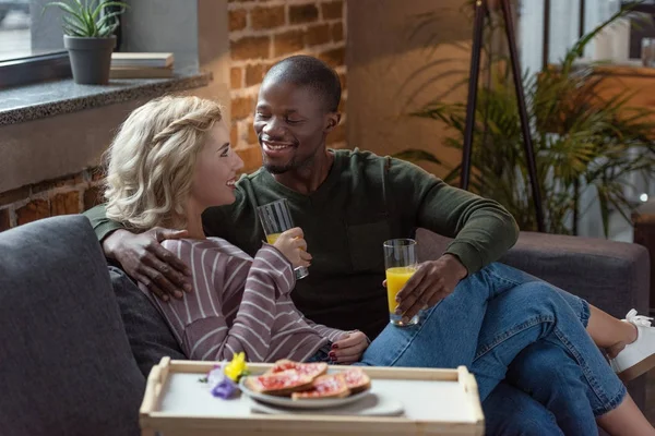 Side view of smiling interracial couple with glasses of juice looking at each other at home — Stock Photo