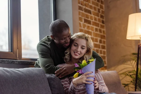 African american man hugging smiling caucasian girlfriend with bouquet of flowers at home — Stock Photo