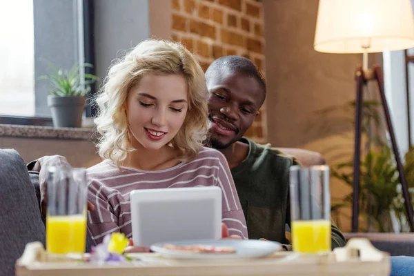 Молодые улыбающиеся многонациональная пара с помощью планшета вместе во время завтрака на дому — стоковое фото