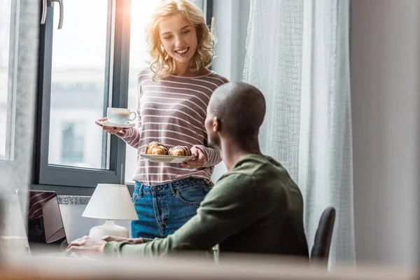 Namorada trouxe xícara de café e croissants para o namorado afro-americano que trabalha em casa — Fotografia de Stock