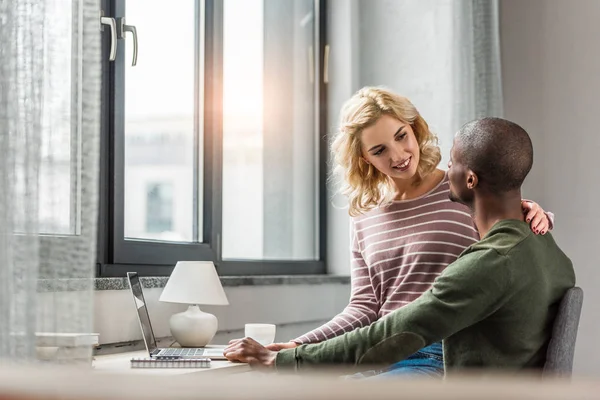 Alegre mujer sentado en afroamericano novios rodillas en mesa con portátil en casa - foto de stock