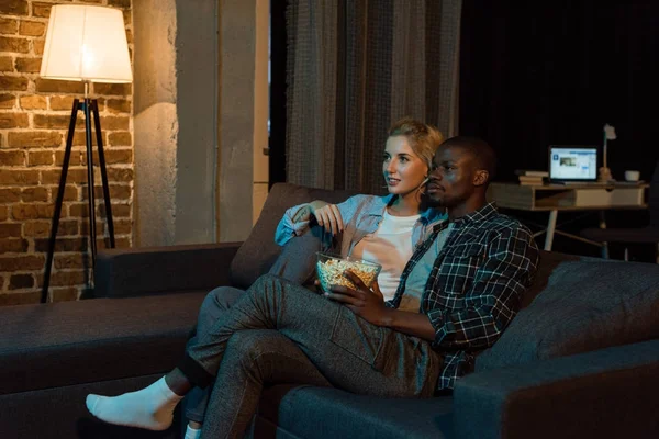 Молода багатоетнічна пара з попкорном дивиться фільм разом на дивані вдома — стокове фото