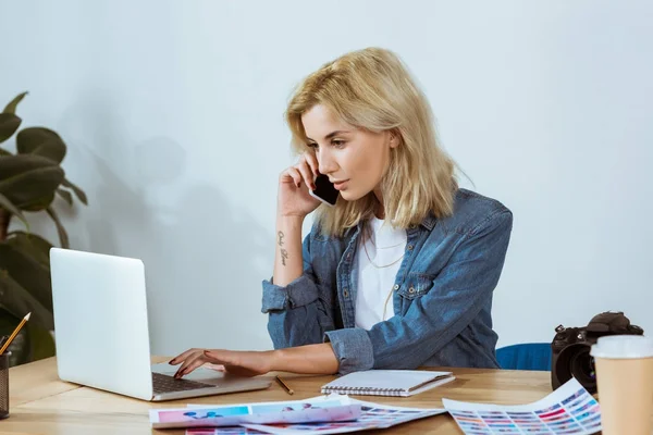 Ritratto di giovane fotografo che parla su smartphone mentre digita sul computer portatile sul posto di lavoro in studio — Foto stock