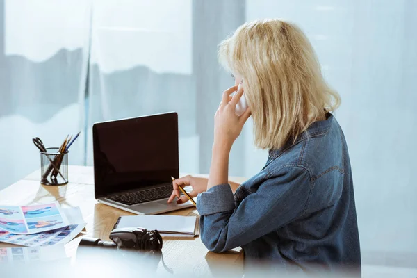 Seitenansicht des Fotografen, der auf dem Smartphone spricht, während er am Arbeitsplatz im Studio am Laptop tippt — Stockfoto