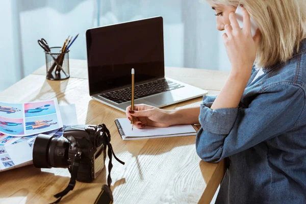 Seitenansicht des Fotografen, der am Arbeitsplatz mit Laptop im Studio auf dem Smartphone spricht — Stockfoto