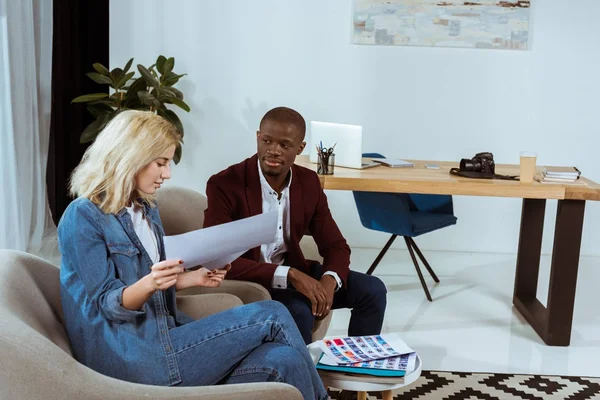 Interracial photographers choosing photos together while sitting in armchairs in office — Stock Photo