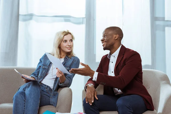 Interracial photographers choosing photos together while sitting in armchairs in office — Stock Photo