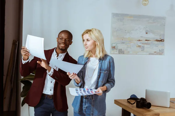 Lächelnde interrassische Fotografen, die Fotos auswählen, während sie im Büro am Fenster stehen — Stockfoto