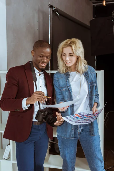 Retrato de fotógrafos multiétnicos sorridentes olhando para o portfólio juntos em estúdio — Fotografia de Stock