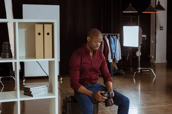 Portrait of african american photographer looking at photo camera in hands while choosing photos in studio — Stock Photo