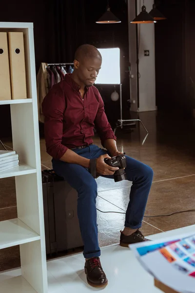 African american photographer looking at photo camera in hands while choosing photos in studio — Stock Photo