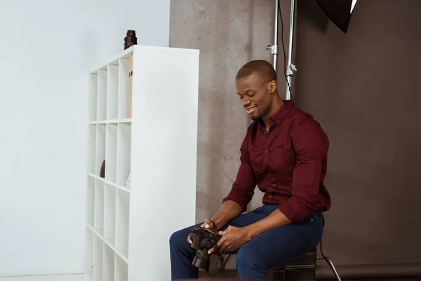 Vista lateral del sonriente fotógrafo afroamericano mirando la cámara fotográfica en las manos mientras elige fotos en el estudio - foto de stock