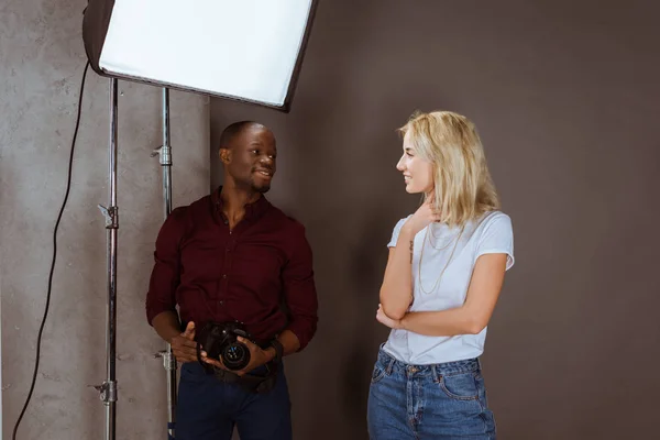 Fotógrafo multiétnico alegre e modelo olhando um para o outro, tendo photoshoot em estúdio — Fotografia de Stock