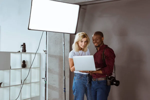 Photographe afro-américain et modèle caucasien choisissant des photos sur ordinateur portable ensemble pendant la séance photo en studio — Photo de stock